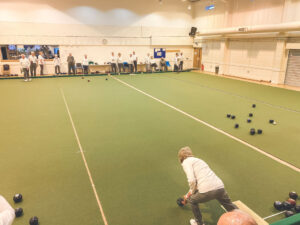 A player bending over to bowl on the indoor carpet, with a wide view of the well-lit facility and other players in the background