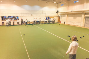 A bowler actively delivering their bowl along the indoor carpet green, with fellow members looking on