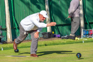 A player in an exaggerated delivery stance on the outdoor green, with the green fencing visible in the background
