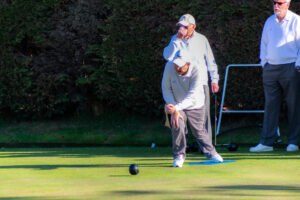 A member in light-coloured clothing in mid-delivery on the outdoor green, with fellow members watching nearby